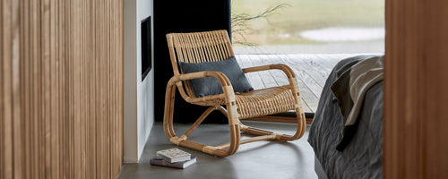 Vævet stol med en behagelig pude, placeret ved siden af en seng, der viser et moderne og stilfuldt design i en fredfyldt indstilling.