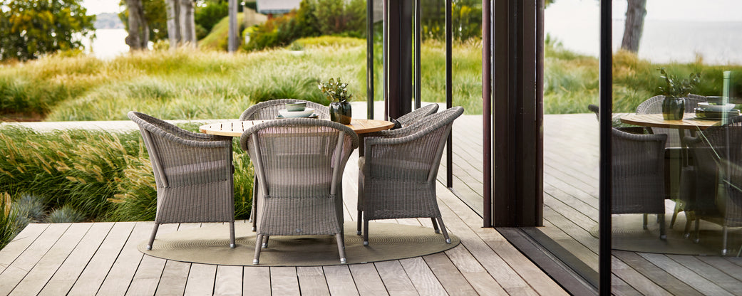Stylish dining setup featuring a round table and woven chairs, surrounded by lush greenery and a modern glass backdrop.