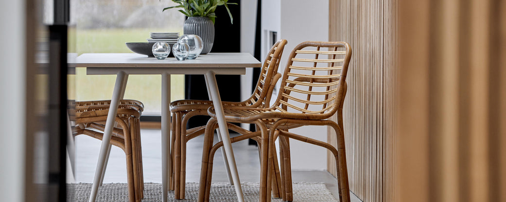 Modern dining table set with a sleek white tabletop and woven chairs, accented by decorative items and greenery.