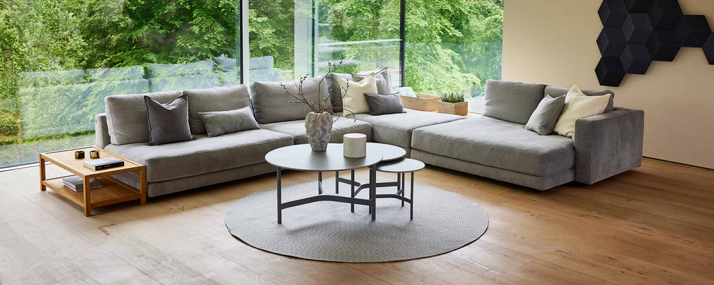 Et rummeligt, moderne opholdsområde med en stor grå sektionalsofa, et rundt tæppe og stilfulde kaffeborde, med grønne områder synlige udenfor.