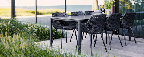 Stylish black dining set with woven chairs, surrounded by lush greenery and a serene view in the background, creating a relaxed vibe.