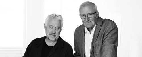 To mænd poserer sammen med venlige udtryk, iført afslappet tøj mod en lys baggrund, der udstråler samarbejde og kreativitet.