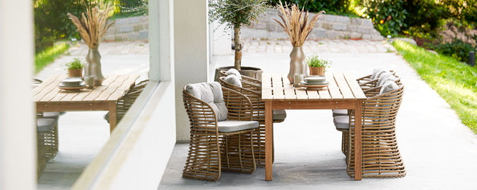 et stilfuldt træbord omgivet af vævede stole, med dekorative planter og grønt i baggrunden, perfekt til selskaber.