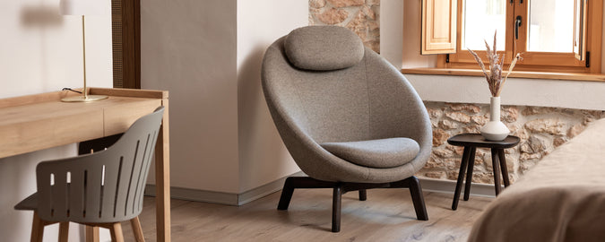 Hyggeligt siddeområde med en moderne stol med en rund design, et lille sidebord og et minimalistisk skrivebord. Naturligt lys strømmer ind gennem et vindue.