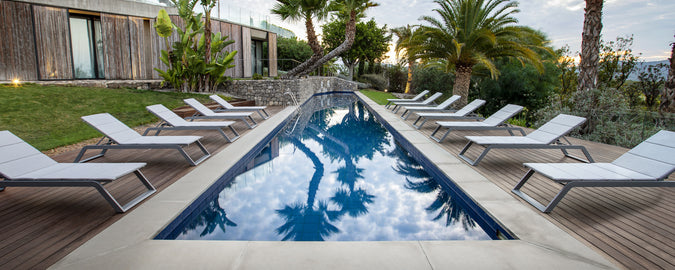 En fredfyldt poolscene med en række hvide solbede ved siden af et roligt vand, omgivet af frodig vegetation og palmetræer.