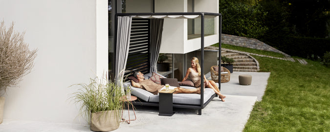 Moderne udendørs loungeområde med en sort daybed med grå puder og hvide gardiner.