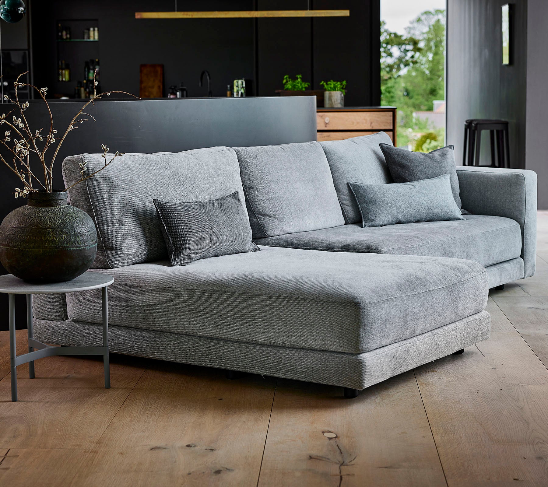 Grå sektionelt sofa med tekstureret stof og dekorative puder, ledsaget af et rundt sidebord og en stilfuld vase.
