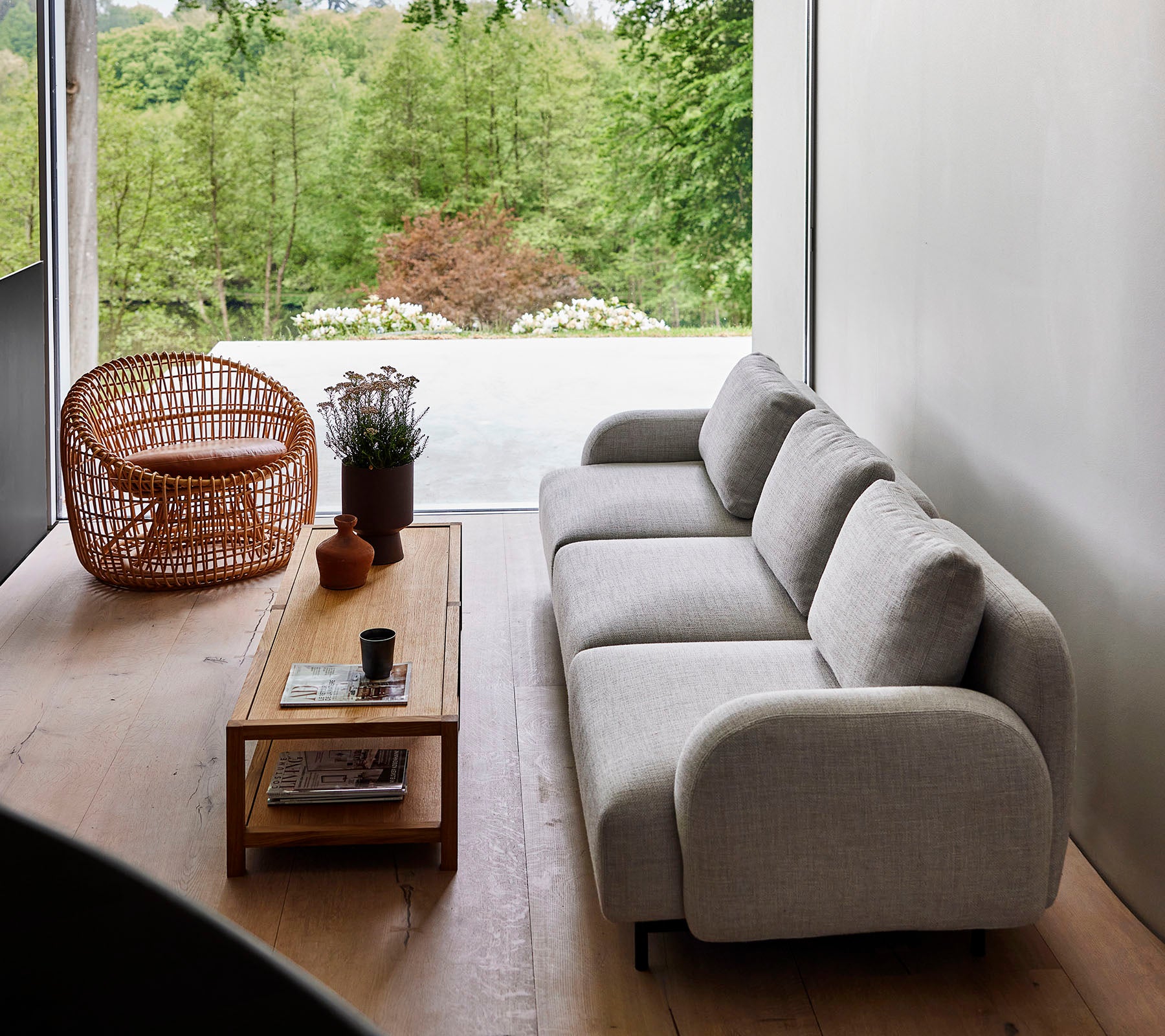 Moderne sofa i lyst stof parret med et trækaffebord og en kurvestol, omgivet af grønne planter. Naturligt sollys forbedrer den rolige atmosfære.