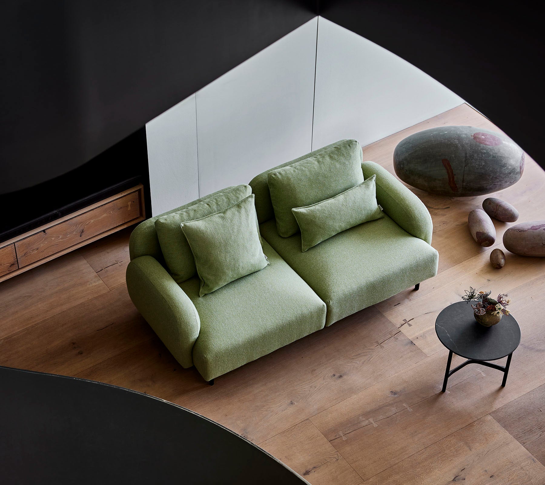 Stilfuld grøn sofa med bløde puder ved et lille rundt bord, sat mod en moderne baggrund med trægulv og glatte sten.