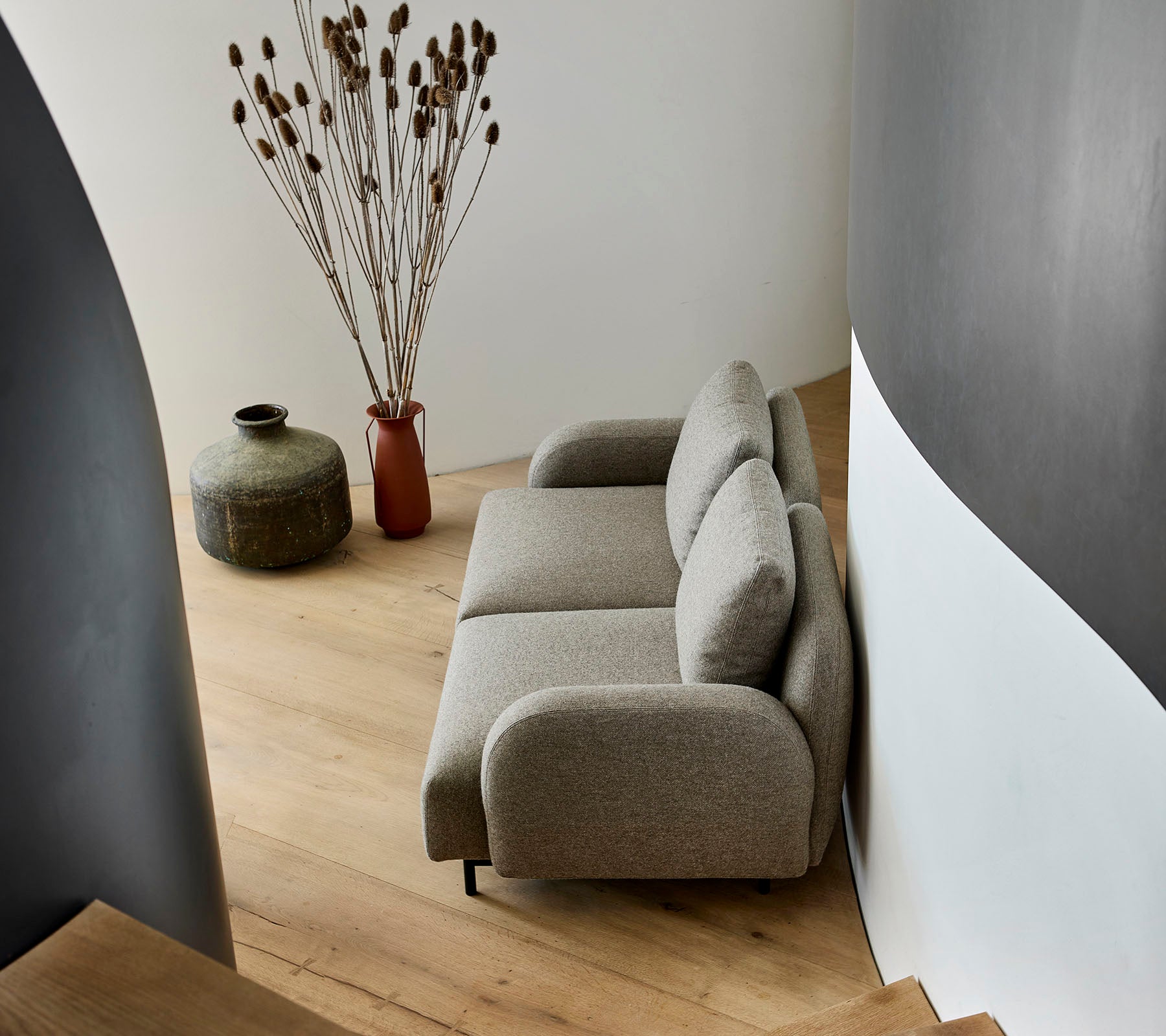 En hyggelig beige sofa placeret ved siden af en dekorativ vase og tørrede planter, mod en buet væg og trægulv.