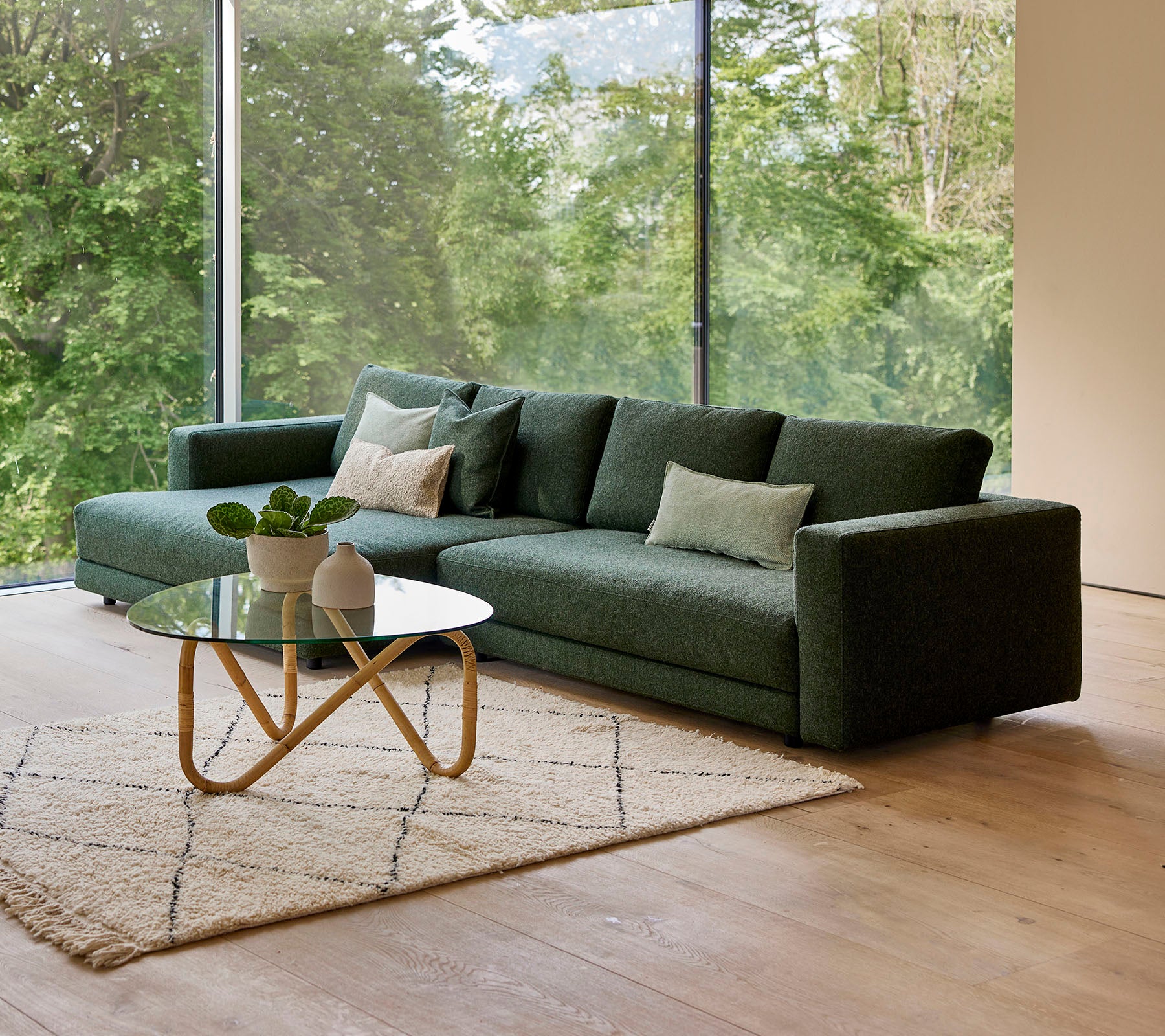 En moderne grøn sofa med puder, akkompagneret af et rundt kaffebord og et dekorativt tæppe, sat mod en baggrund af grønne planter.