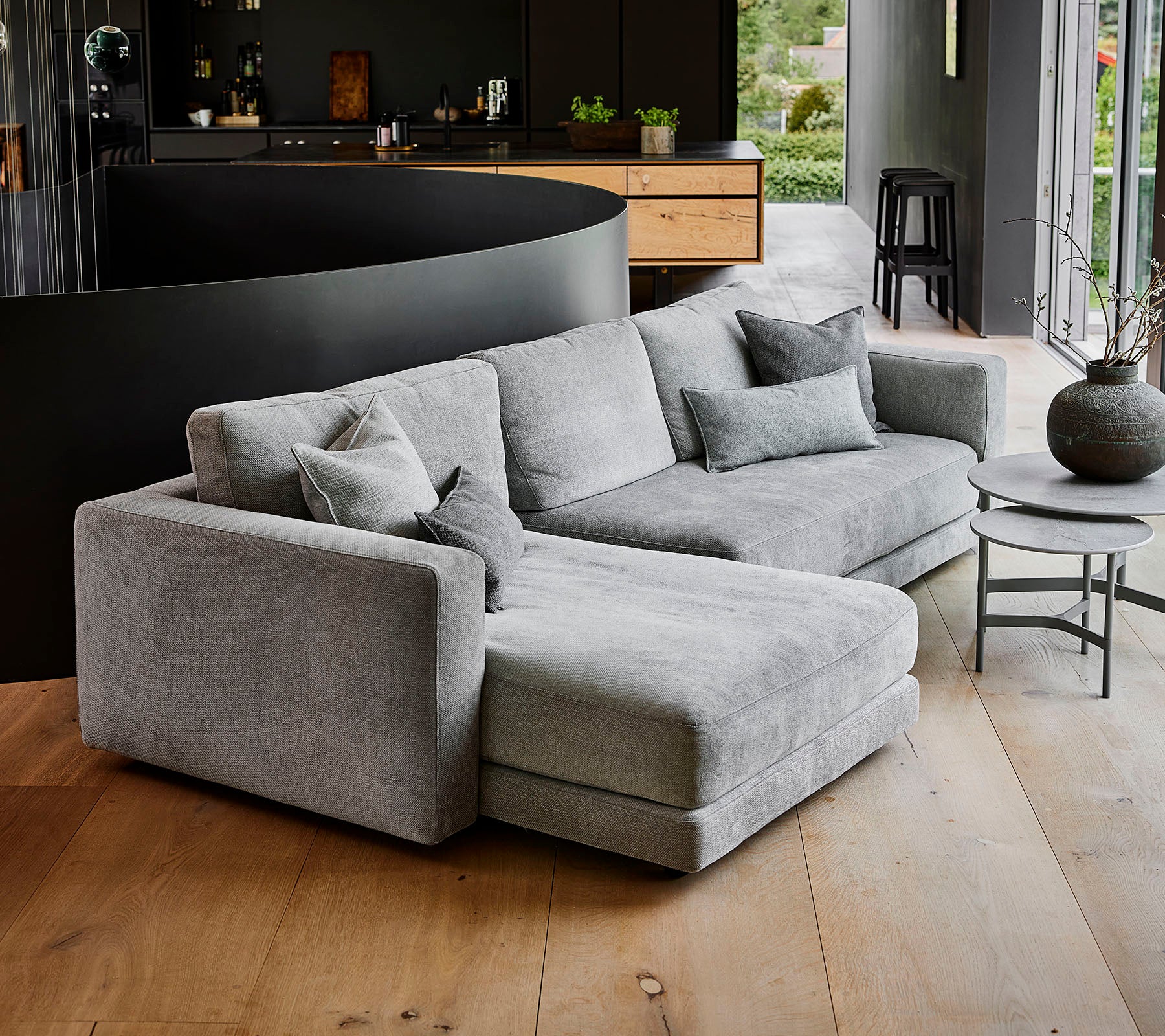 Moderne grå sektion sofa med mange puder, ledsaget af et rundt sofabord og en dekorativ vase. Simpelt, elegant design.