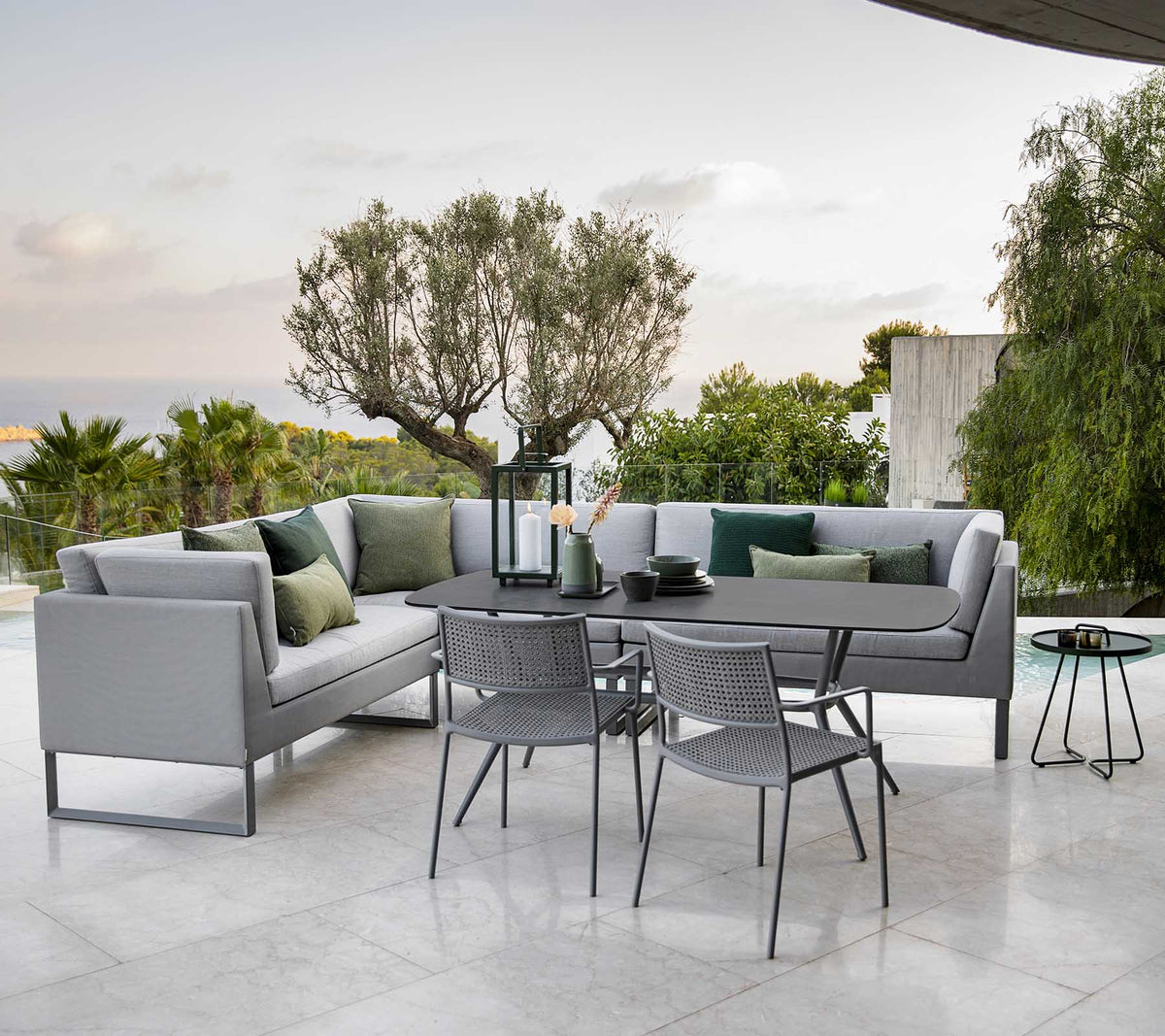 Moderne udendørs siddeområde med en stilfuld sektion sofa, et spisebord og metalstole omgivet af grønne planter.