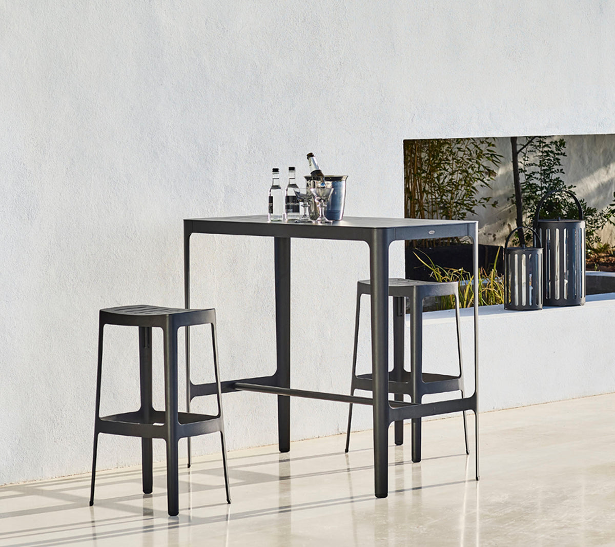 Et slankt bord sat med to matchende taburetter, der har et minimalistisk design og flasker placeret ovenpå, mod en neutral baggrund.