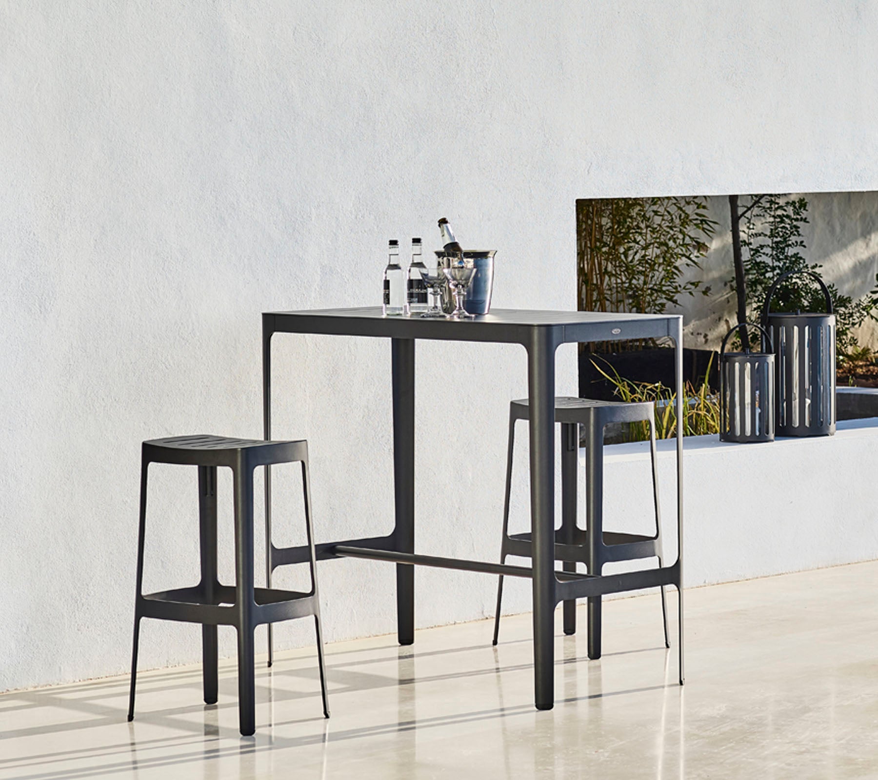 Et slankt bord sat med to matchende taburetter, der har et minimalistisk design og flasker placeret ovenpå, mod en neutral baggrund.