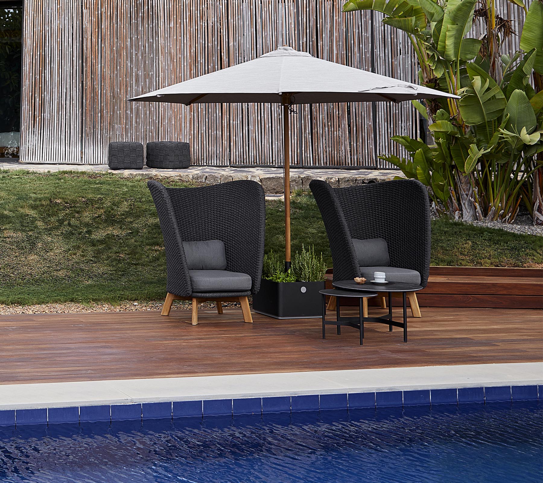 To stilfulde havestole med puder under en stor parasol, ledsaget af et lille bord, ved siden af en swimmingpool.