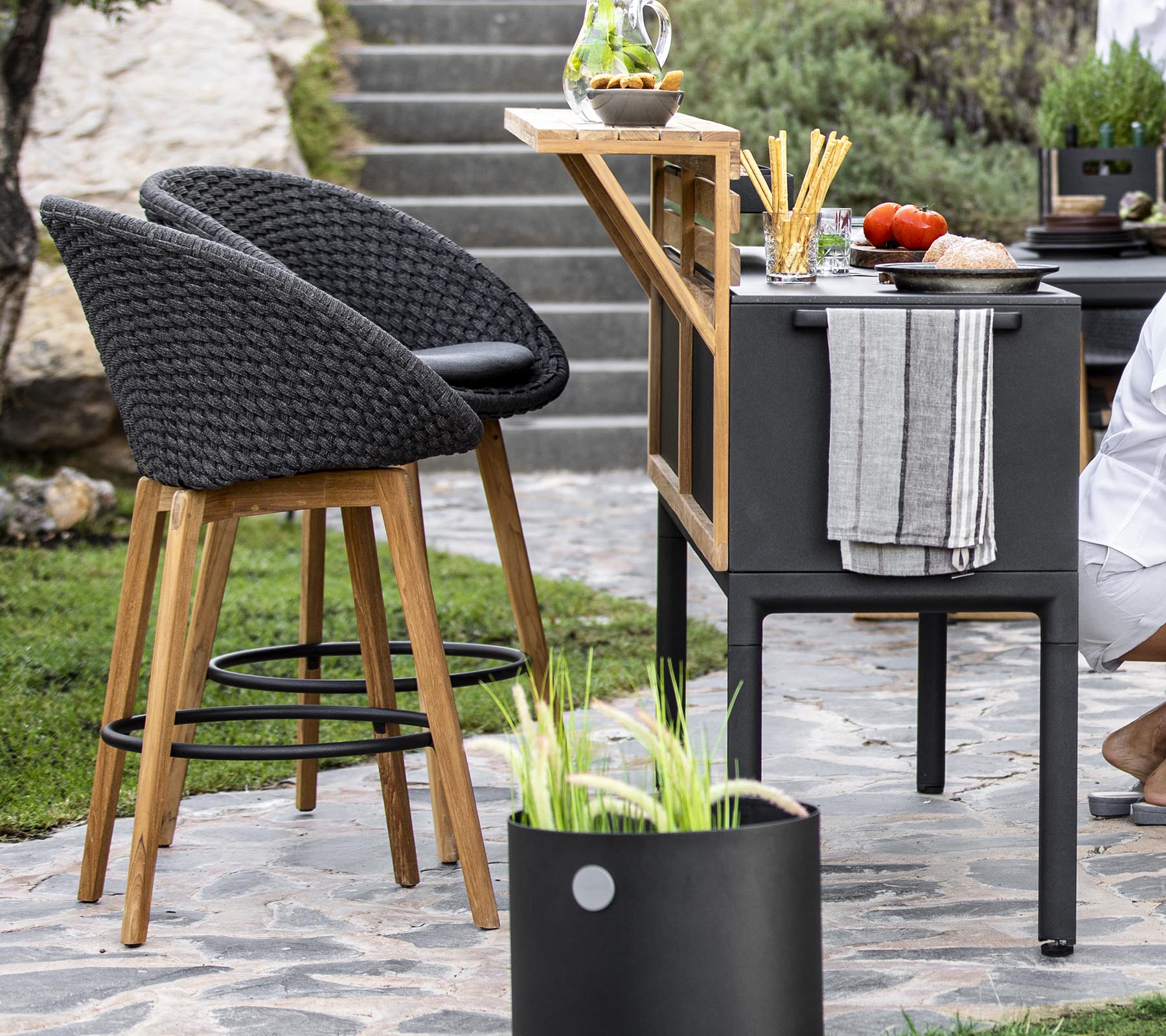 Elegant udendørs siddepladser med rattan stole, en moderne barvogn og friske ingredienser arrangeret på en bordplade, suppleret med dekorative planter.