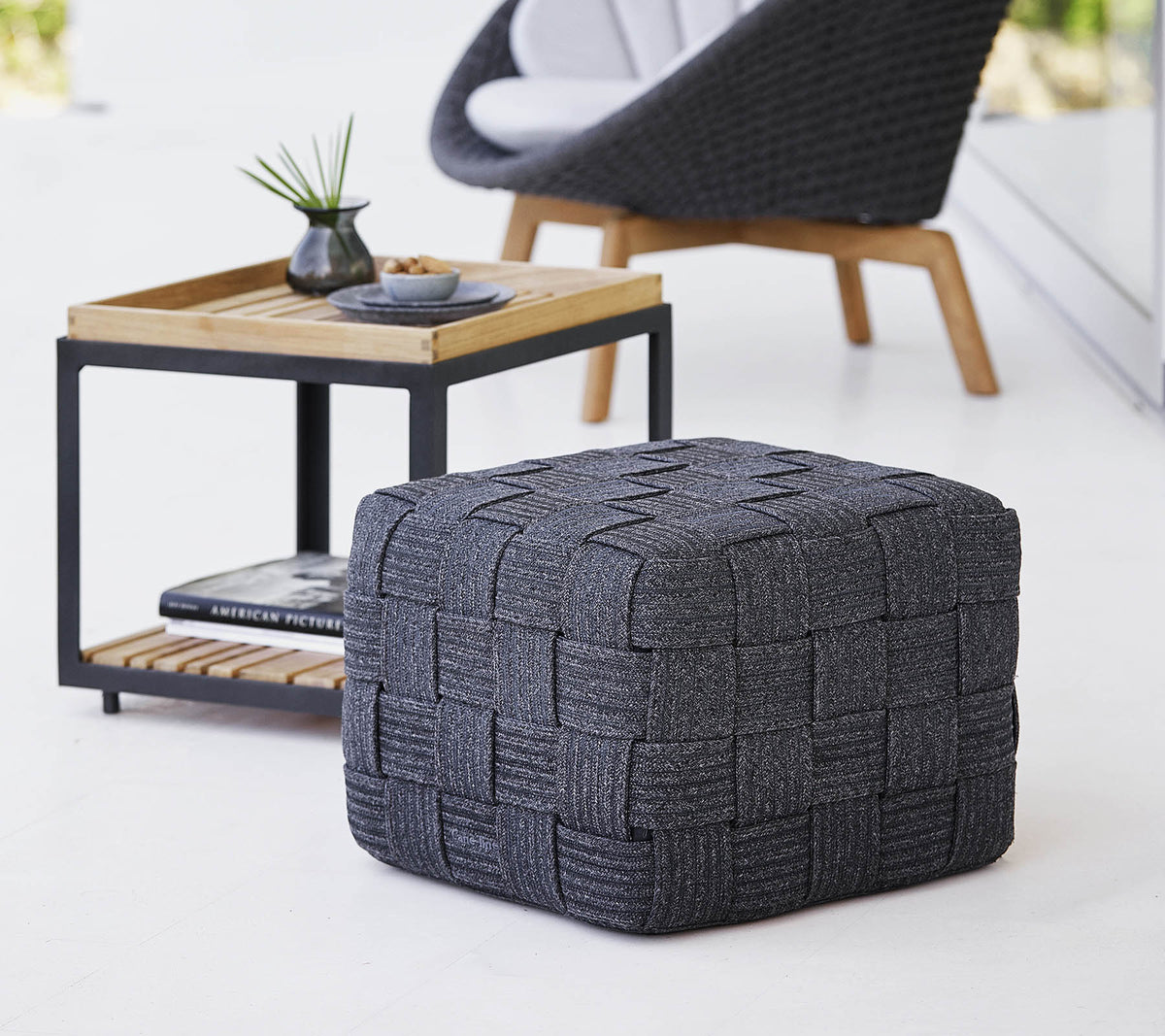 En struktureret grå vævet pouf placeret nær et træsidebord med et minimalistisk design, som indeholder små dekorationsgenstande.