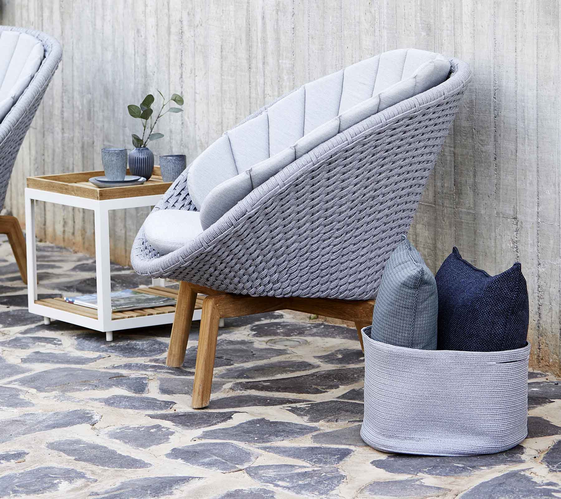 Moderne grå lounge stol med et vævet design, ledsaget af puder og et lille træsidebord. Stengulvet forbedrer æstetikken.