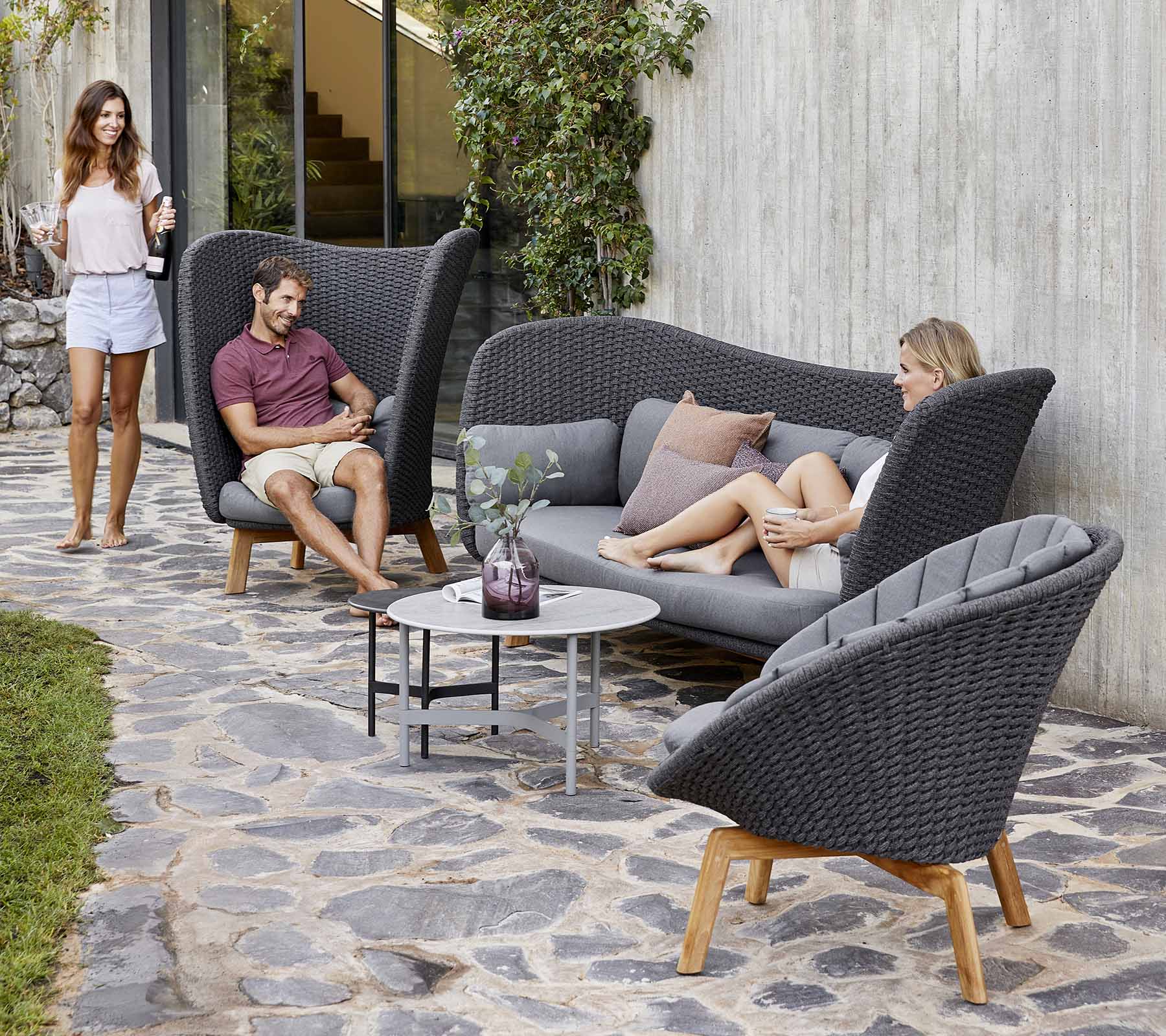 Stilfuld udendørs siddearrangement med en buet sofa og stole, med et lille bord, omgivet af grønne planter.