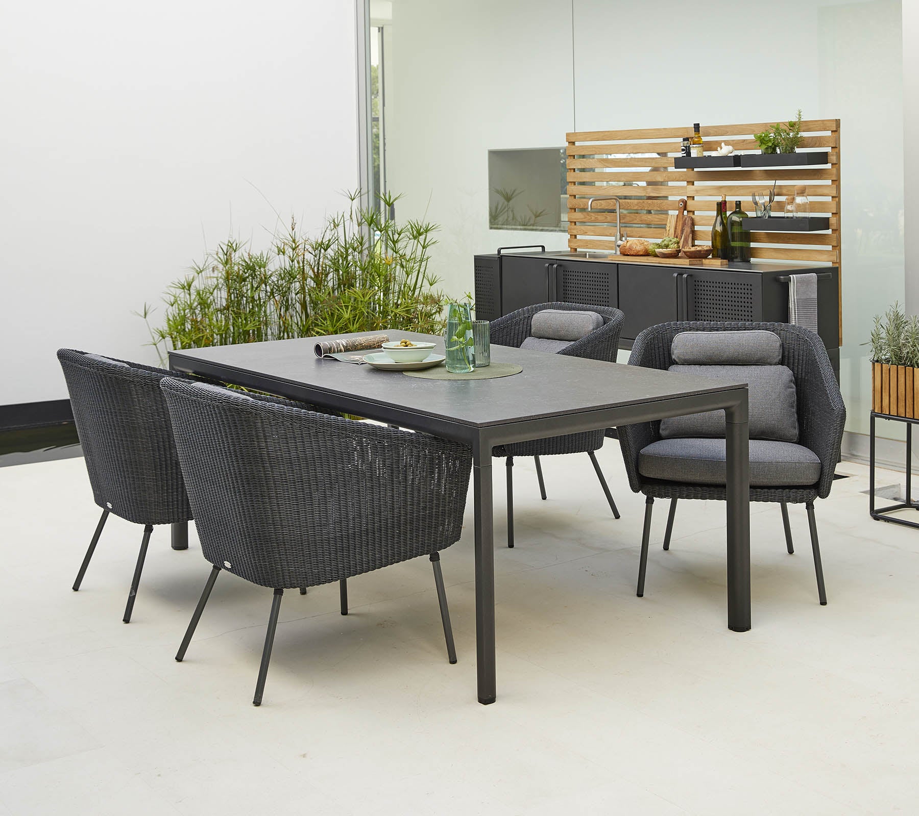 Moderne udendørs spisebordssæt med et slankt bord og fire stilfulde stole med komfortable puder, omgivet af grønne planter.
