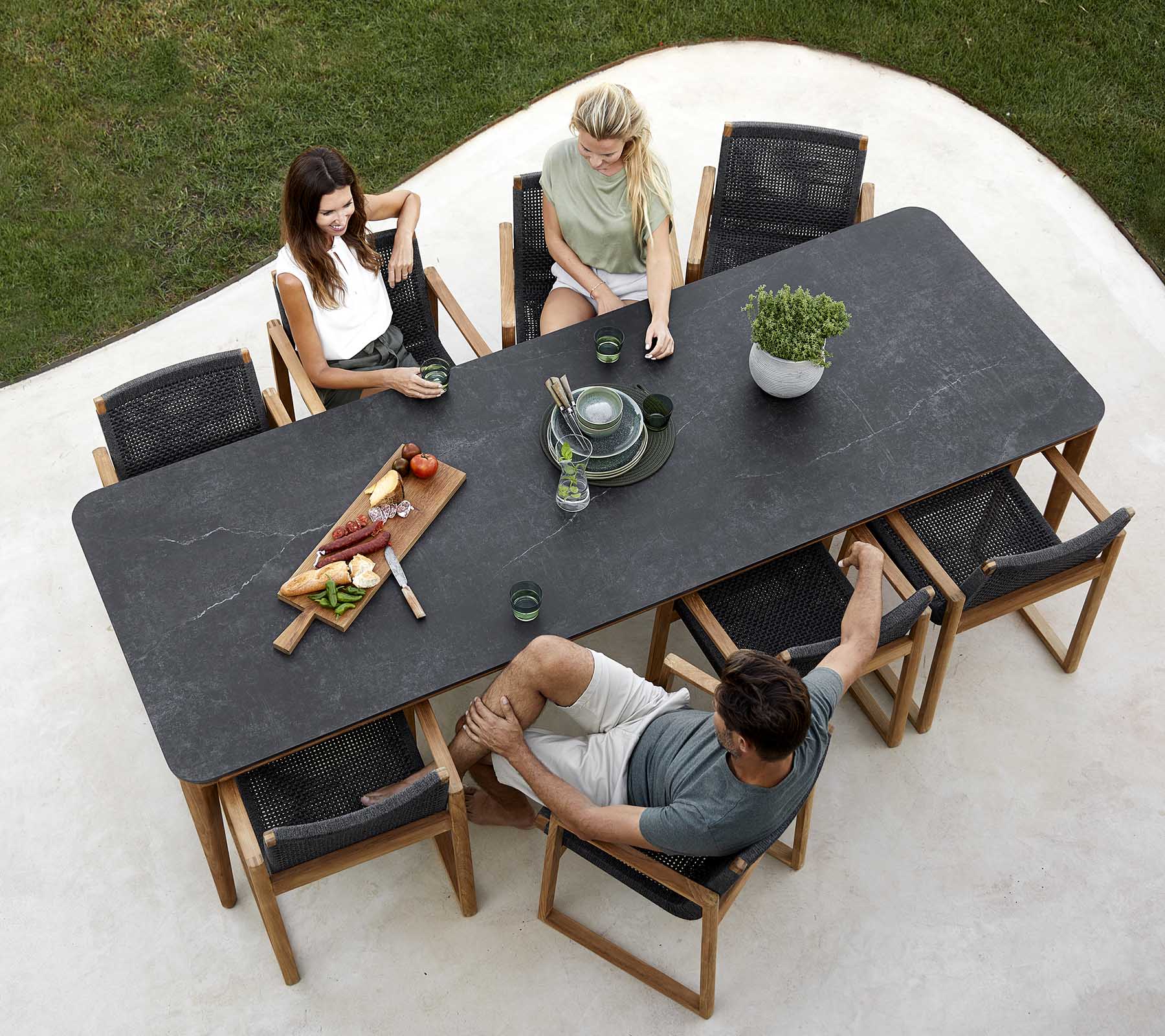En gruppe mennesker nyder et måltid omkring et stort bord med retter, drikkevarer og en plante-centerpiece. Uformel spise-atmosfære.