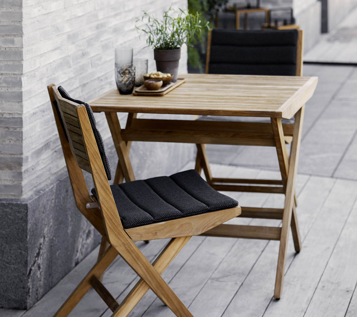 Sæt af træbord og stole med sorte puder, prydet med potteplanter og dekorativt glas. Perfekt til en hyggelig atmosfære.