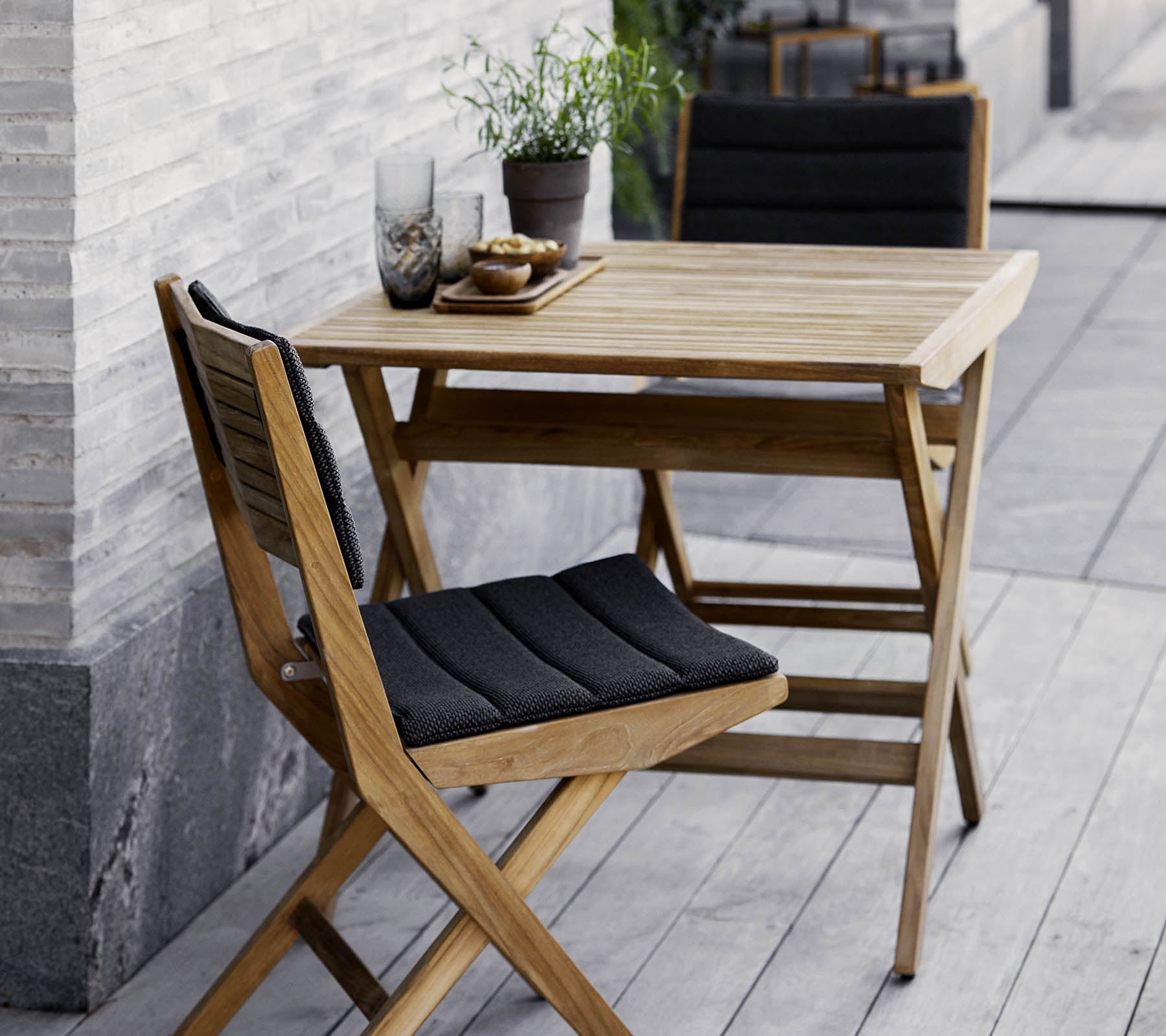 Sæt af træbord og stole med sorte puder, prydet med potteplanter og dekorativt glas. Perfekt til en hyggelig atmosfære.