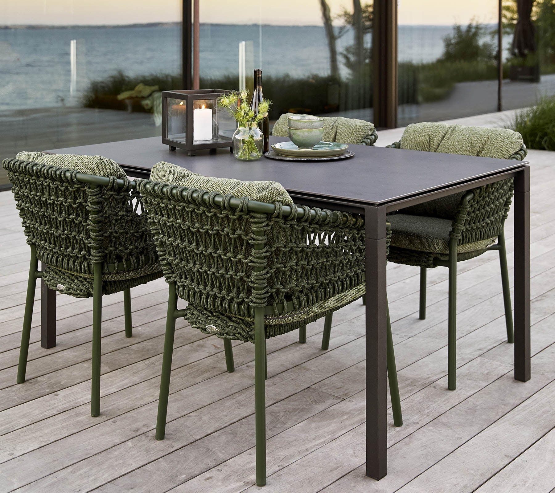 Stylisk udendørs spisestel med et rektangulært bord og fire polstrede, vævede stole, suppleret med et blomsterarrangement i midten.