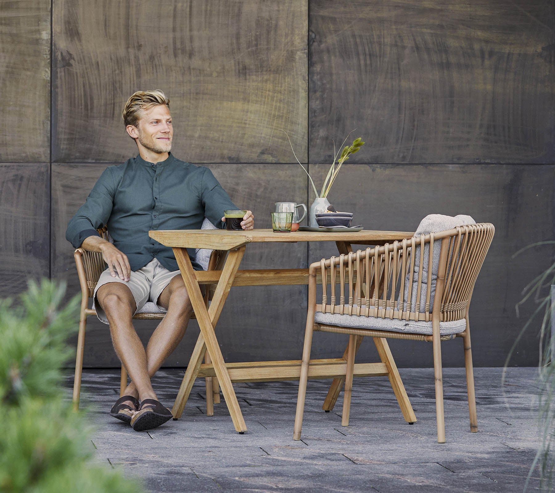 En mand sidder afslappet ved et stilfuldt trætabel med en let drink, omgivet af grønne planter og en struktureret baggrund.