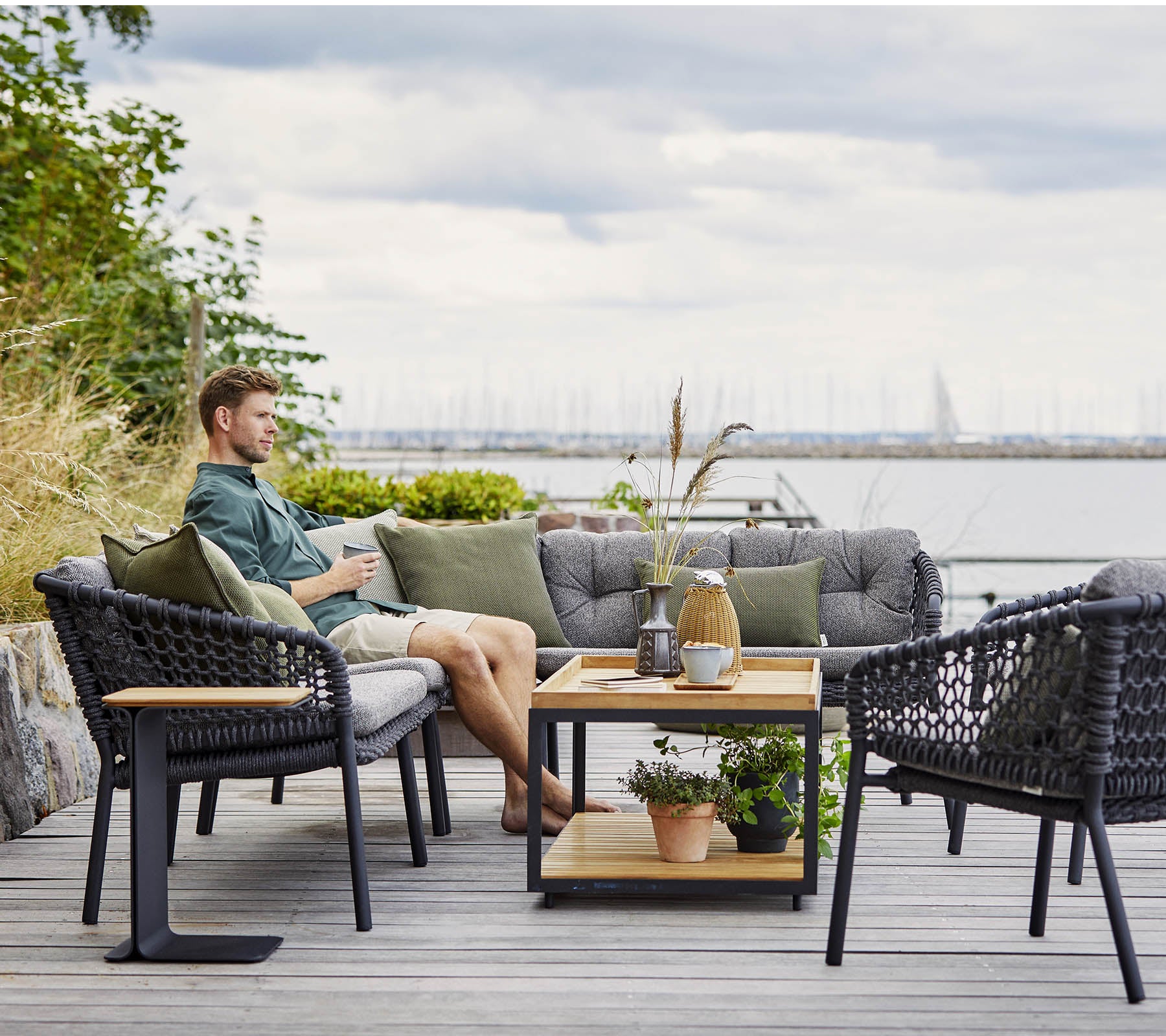 Et komfortabelt siddeområde med en mand, der slapper af, med stilfulde rattanlænestole og et trækaffebord udsmykket med planter og dekorative genstande.