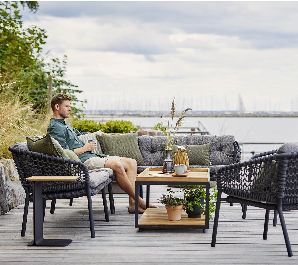 Et komfortabelt siddeområde med en mand, der slapper af, med stilfulde rattanlænestole og et trækaffebord udsmykket med planter og dekorative genstande.