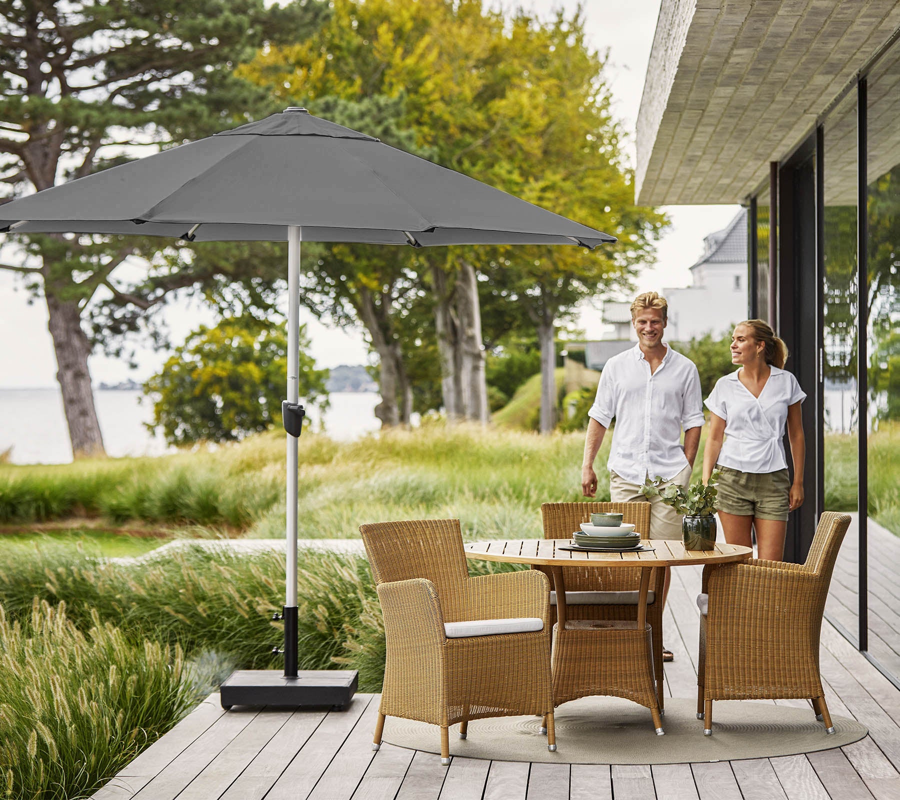 Par, der går på en træterrasse med et rundt bord og flettede stole, skygget af et stort gråt parasol, med udsigt over frodig grøn vegetation.