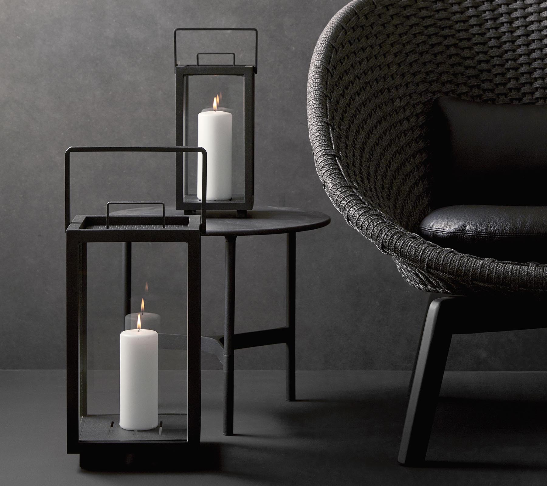 To stilfulde lysekrone med hvide stearinlys, ledsaget af en moderne, tekstureret stol på et minimalistisk bord mod en mørk baggrund.