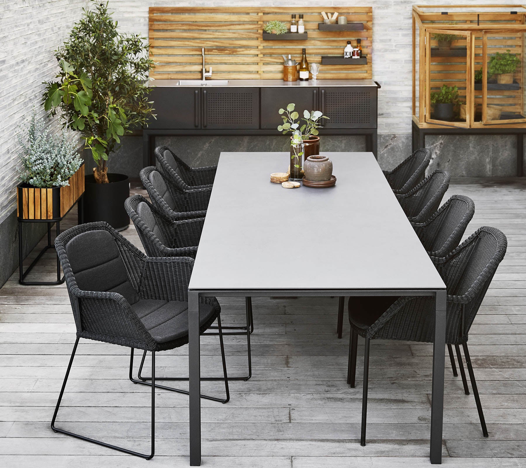 Et moderne spisestueopsætning med et langt bord omgivet af elegante sorte stole, suppleret med dekorative planter og en træbaggrund.