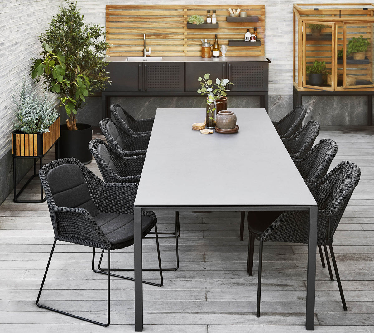 Et moderne spisestueopsætning med et langt bord omgivet af elegante sorte stole, suppleret med dekorative planter og en træbaggrund.