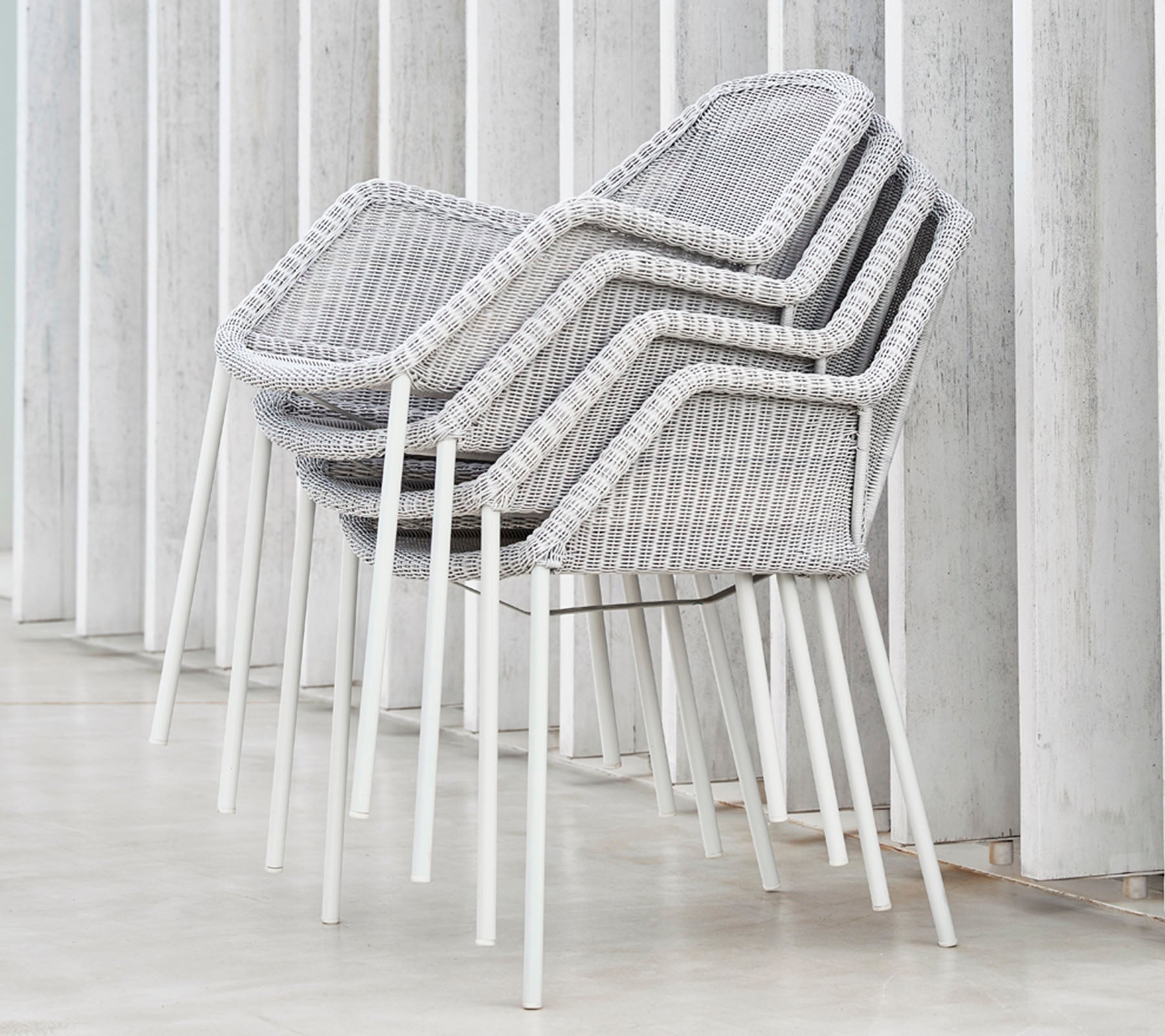 Sammenstillede moderne stole med et vævet design i grå og hvid, hvilende mod vertikale overflader, der viser en slank og minimalistisk æstetik.