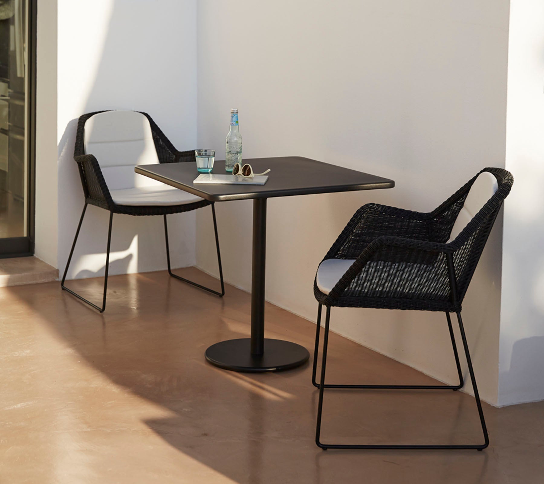 Moderne bord med to stilfulde sorte stole, ledsaget af en vandflaske og solbriller, sat mod en minimalistisk baggrund.
