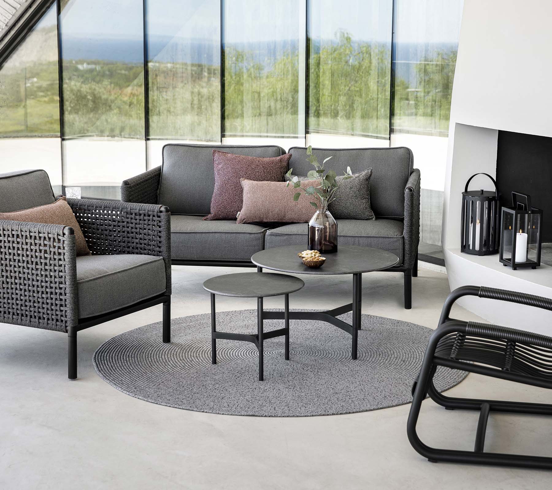 Stilfuld siddeområde med en grå sofa med puder, rundt kaffebord og accentstole på et rundt tæppe. Naturligt lys forbedrer stemningen.