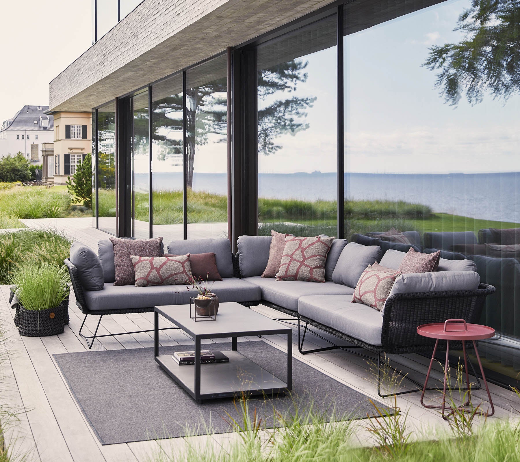 Moderne udendørs siddeområde med en sektionel sofa med puder, et kaffebord og dekorative elementer blandt frodig grøn beplantning.