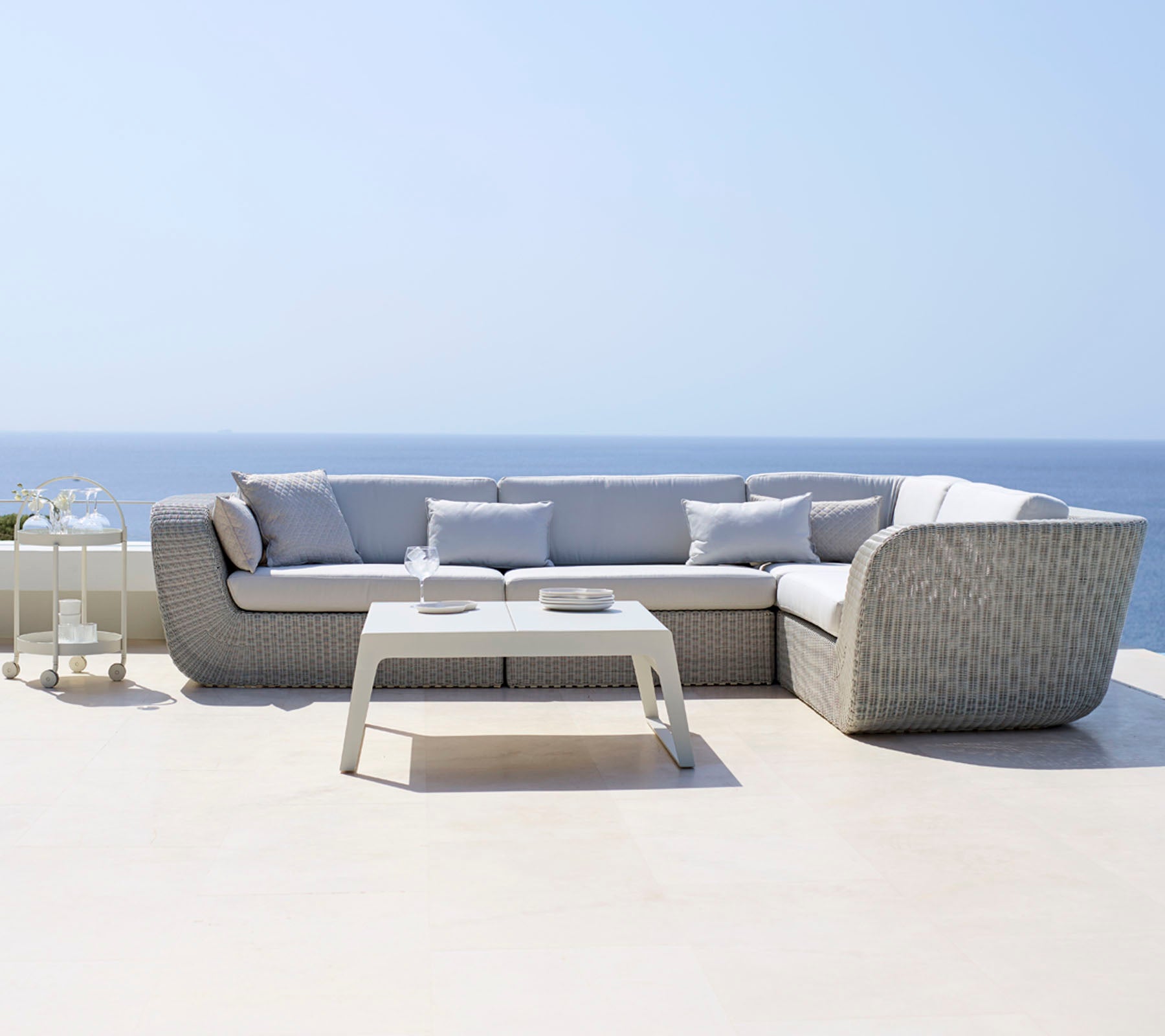 Moderne sektionel sofa med lysegrå puder kombineret med et slankt hvidt sofabord, placeret mod en rolig havudsigt.