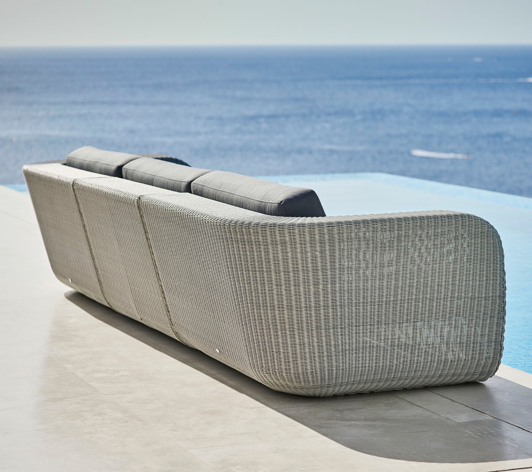 Stilfuld vævet sofa med buet design og bløde puder, der vender ud mod en rolig havudsigt. Ideel til afslapning og udendørs omgivelser.