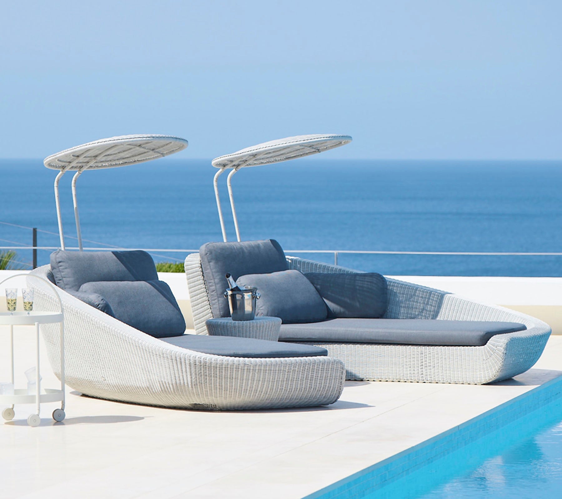 Stilfulde udendørs lounge stole med canopy ved en fredfyldt pool, med udsigt over et roligt hav. Perfekt til afslapning og fritid.