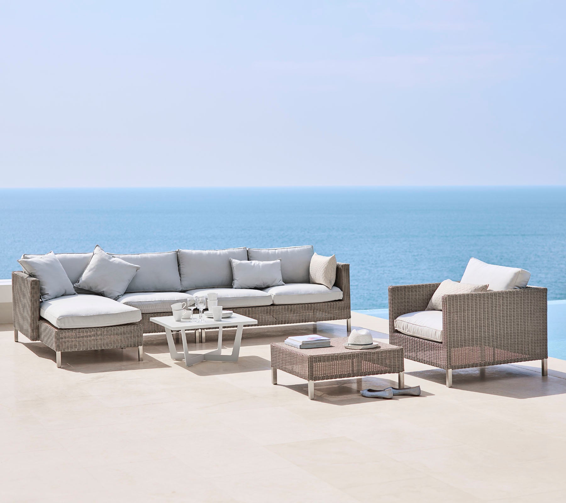 Et stilfuldt udendørs siddearrangement med en stor sofa, en lænestol og et sofabord, alle med lyse puder og flettet materiale, ved havet.