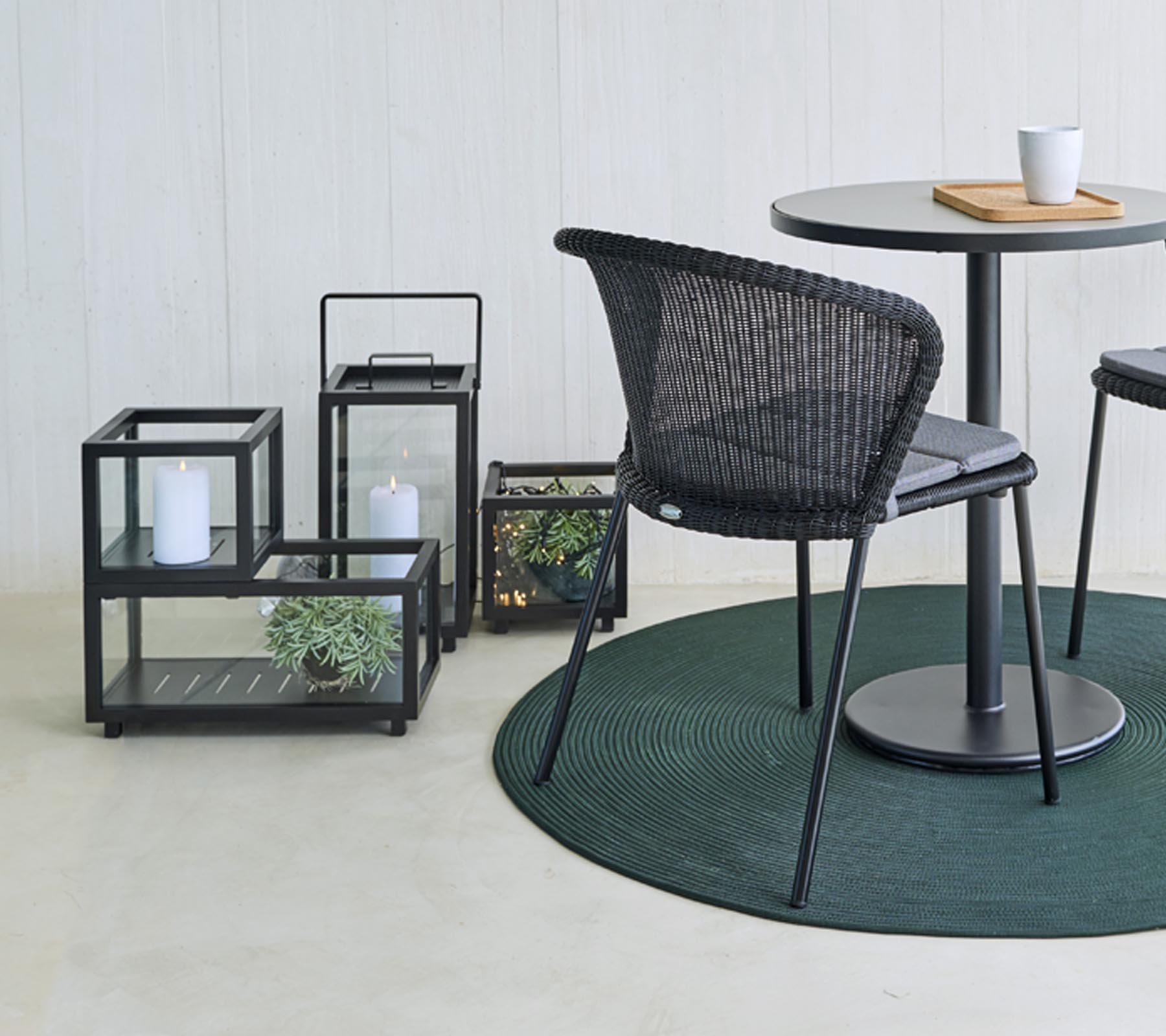 Moderne siddepladser og dekorative glaslanterner med planter, arrangeret omkring et rundt bord, på et rundt grønt tæppe.