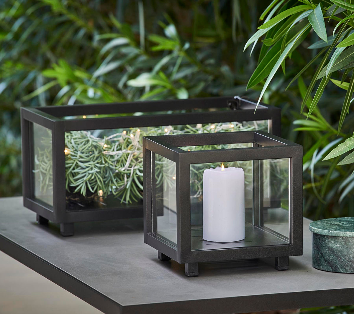 To stilfulde lanterner med glaspaneler, den ene med et hvidt lys og den anden med grønne planter, placeret på et bord midt i frodig løv.
