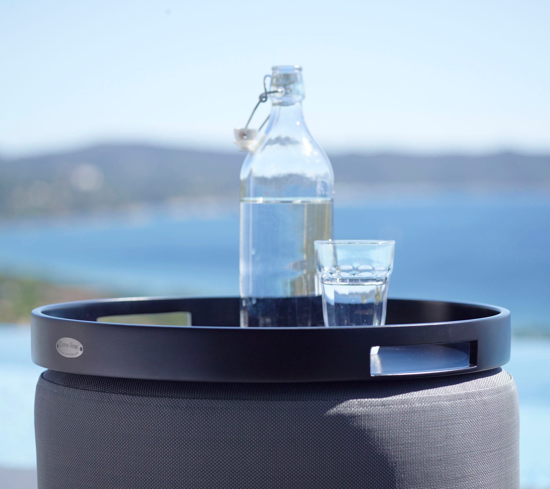 En sort rund bakke holder et glas vand og en glasflaske, sat mod en naturskøn baggrund af havet og fjerne bakker.