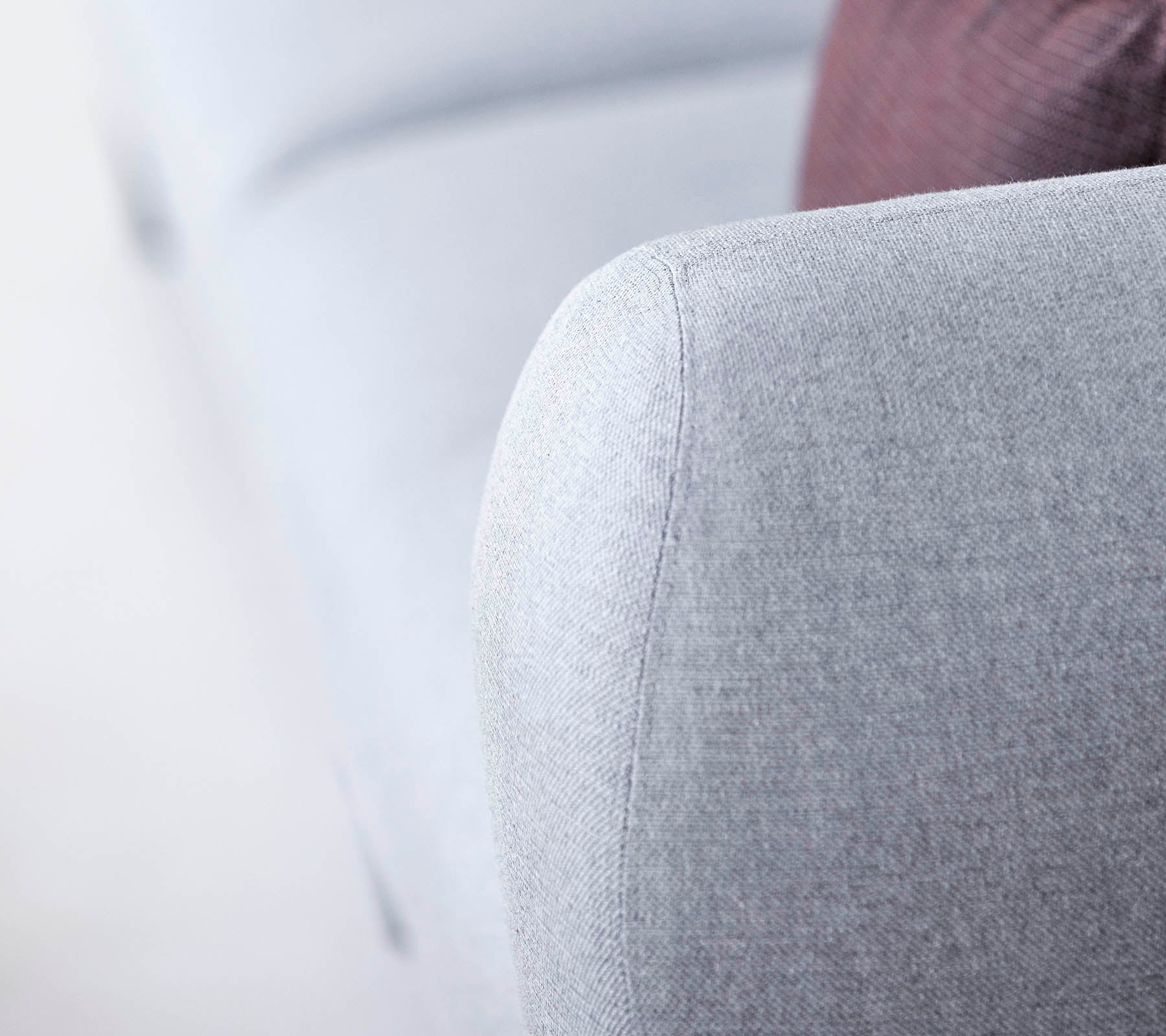 Nærbillede af et lys gråt sofa-arm, der har et tekstureret stof og et blødt, afrundet design, med et glimt af en mørkere pude i baggrunden.