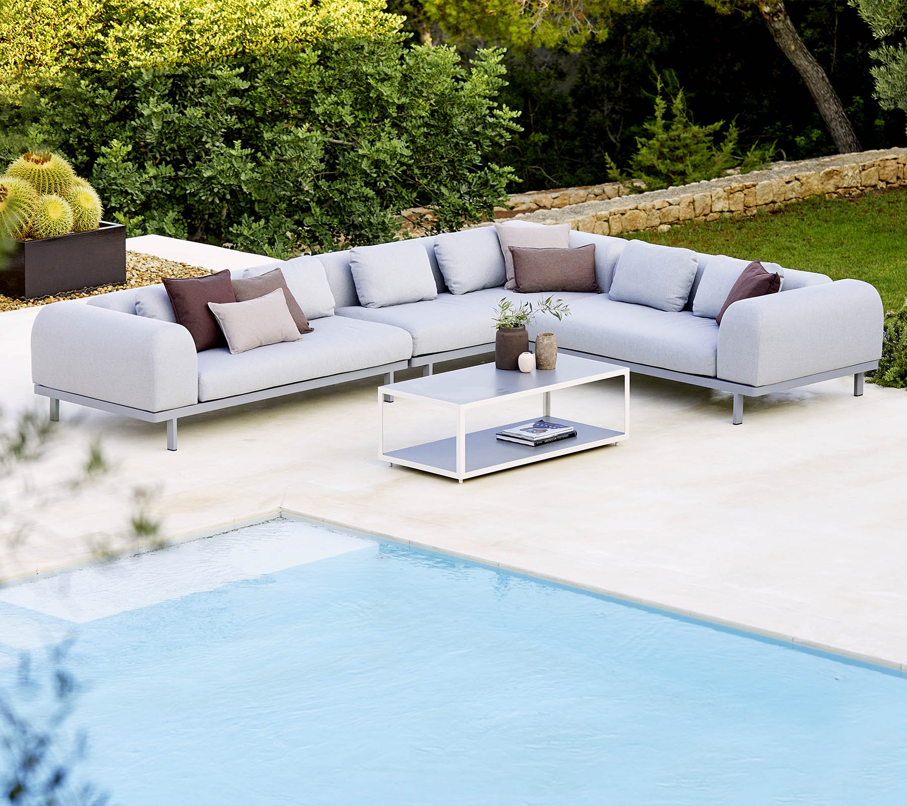 Lys grå sektionel sofa med dekorative puder og et glastable, placeret ved siden af en swimmingpool og frodig vegetation.