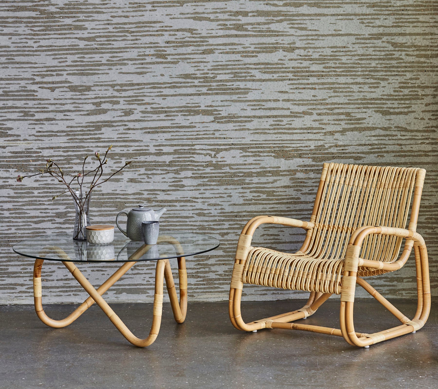 En stilfuld rattan stol ved siden af et glas kaffebord, med en vase med grene og en lille tekande, sat mod en tekstureret baggrund.